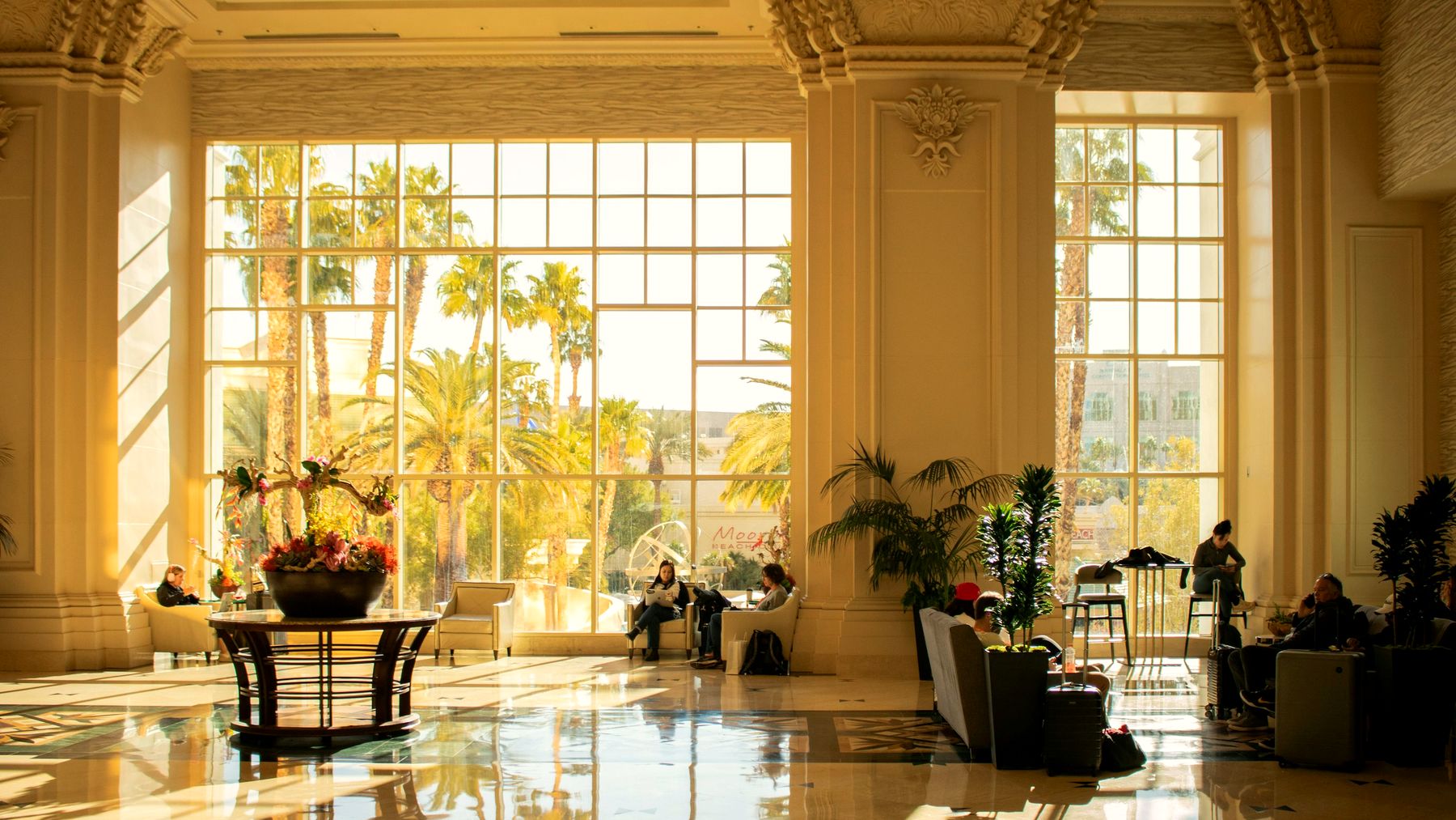 A grand hotel lobby in warm afternoon light with tall windows and palms